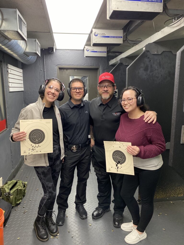 Two Colorado Tactics instructors with two students at an indoor range, the students holding up their shot targets. All four wearing eye and ear protection.