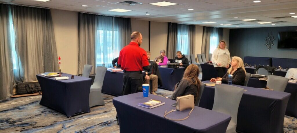 An instructor teaching a seated class of students in a conference room, course materials on the tables and a training pistol at the front.