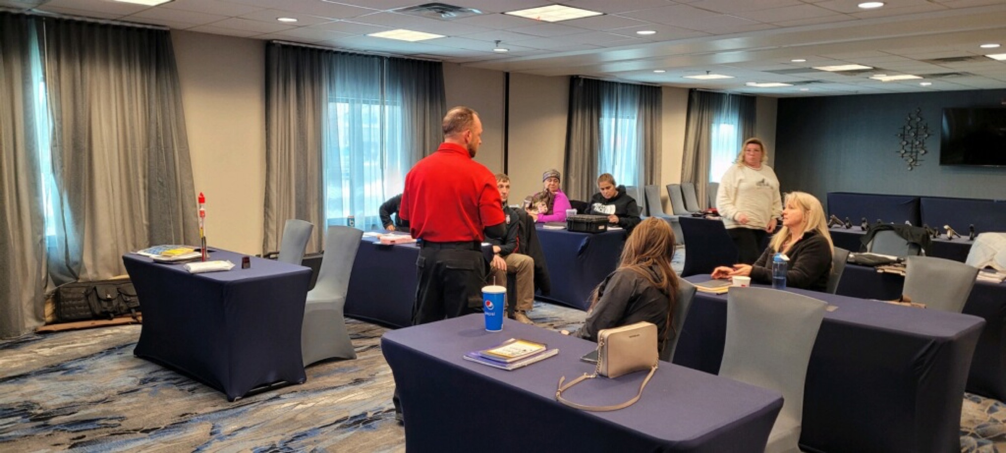 An instructor teaching a seated class of students in a conference room, course materials on the tables and a training pistol at the front.