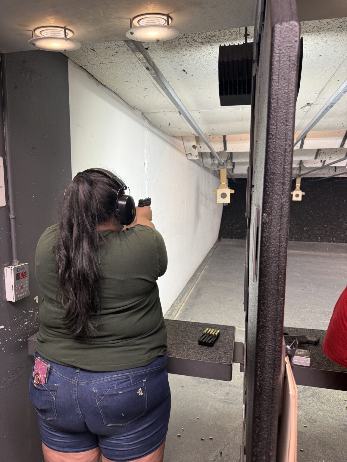 A student in ear defenders firing a pistol down an indoor range lane, spare magazines and ammunition laid out on the bench.