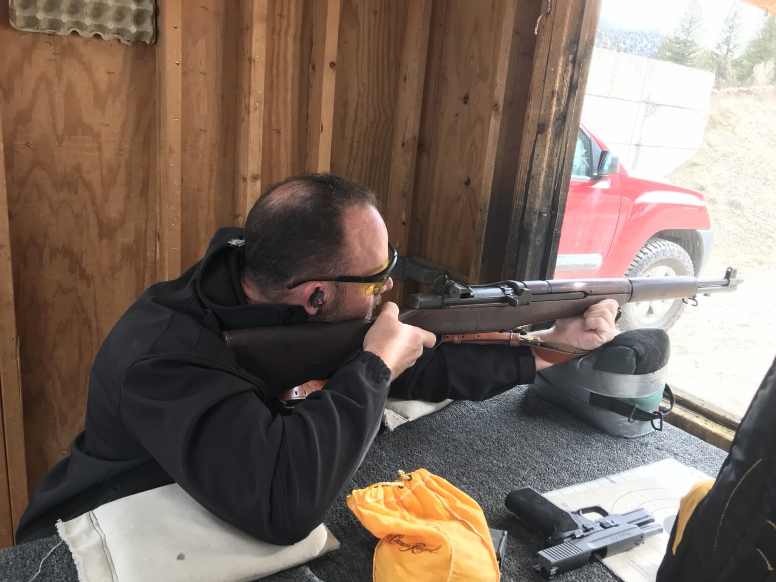 A shooter in safety glasses and ear plugs firing a rifle from a sandbag rest at a covered outdoor range.