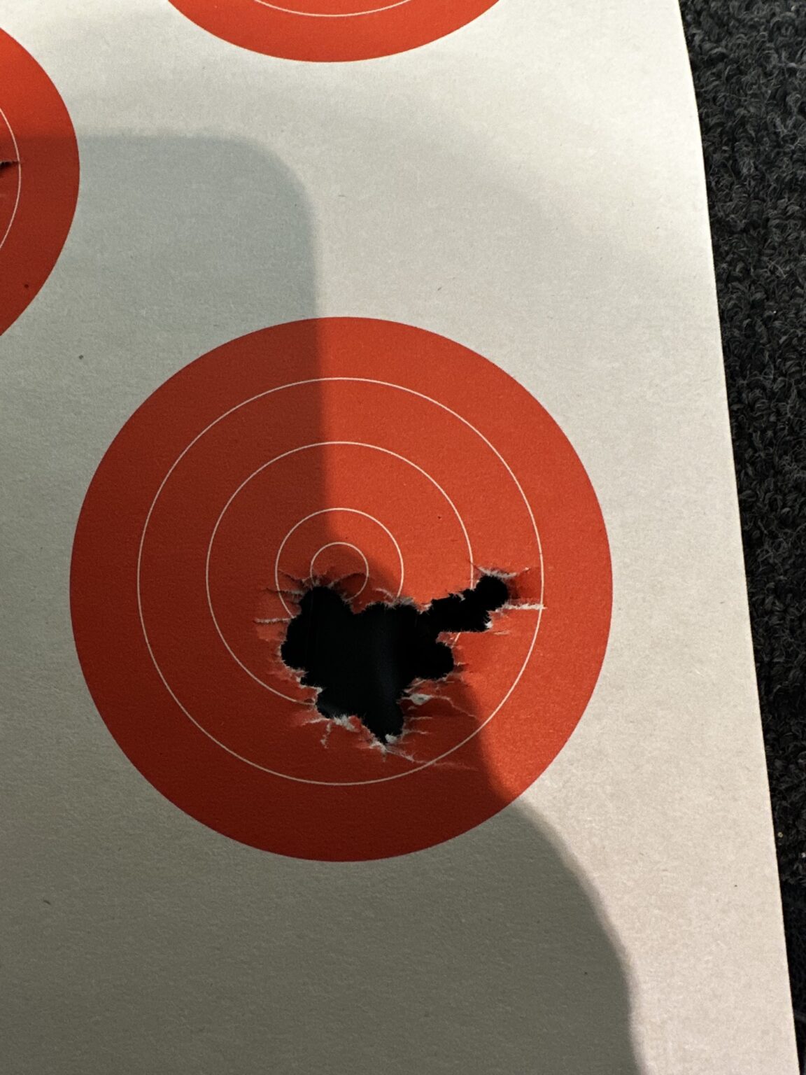 Close-up of a paper target where repeated shots have torn one ragged hole through the center of the orange bullseye.
