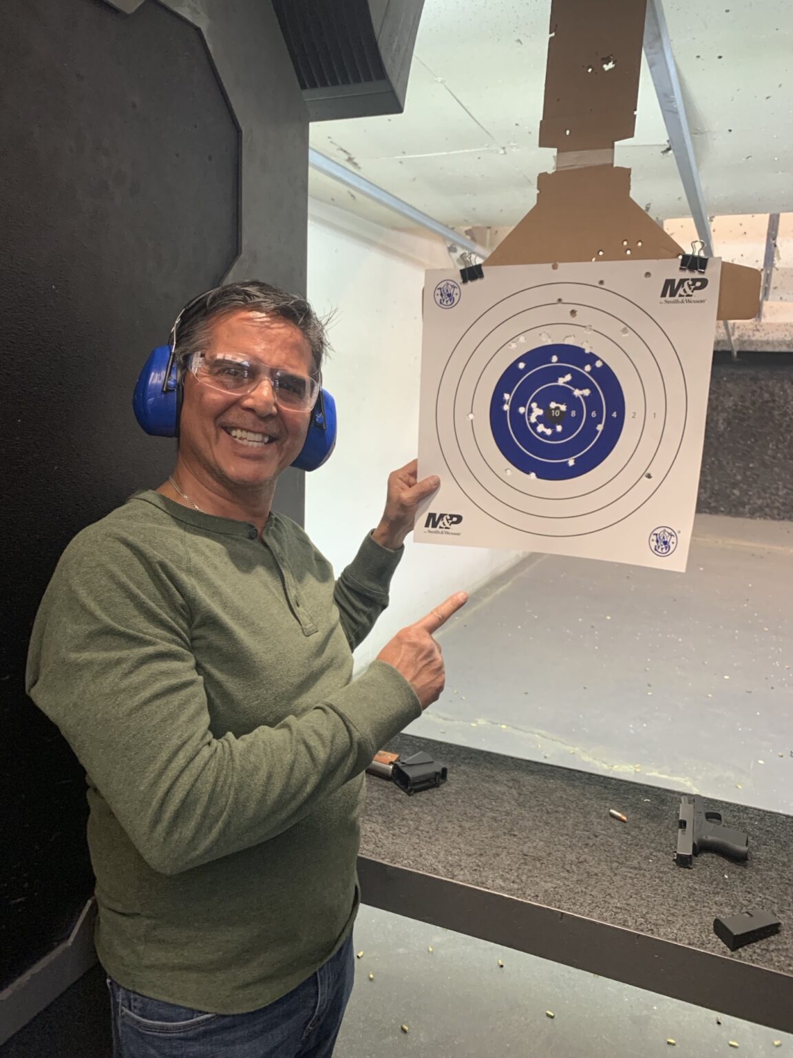A student in eye and ear protection smiling and pointing at his paper target, shots grouped tightly in the center ring, at an indoor range.